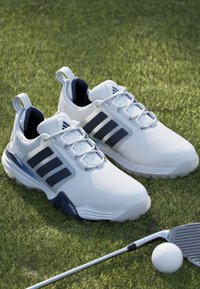 Λευκά παπούτσια του γκολφ με μαύρες ρίγες στο χορτάρι, συνοδευόμενα από ένα γκολφ μπαστούνι και μπάλα.