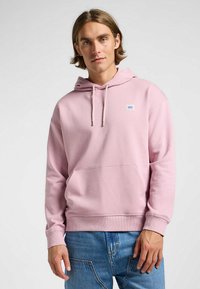 Jasnoróżowa bluza z kapturem, z troczkami i kieszenią kangurką. Posiada małą naszywkę z logo na piersi. Wykonana z miękkiego materiału, o luźnym kroju, z długimi rękawami.