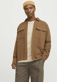 Brown button-up shirt with two chest pockets, worn over a light beige knit sweater, paired with gray trousers, smooth fabric texture.