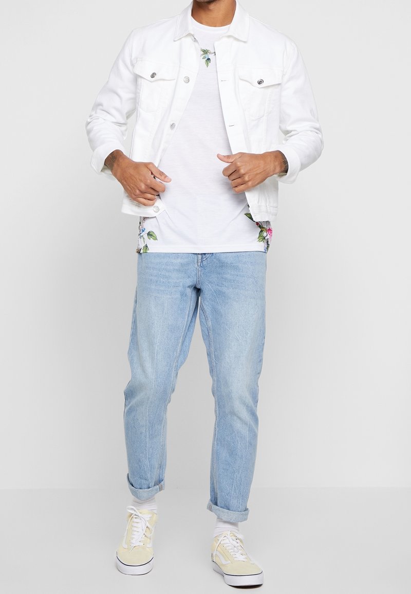 Homme portant une veste en jean blanche par-dessus un t-shirt blanc à fleurs, un jean bleu clair avec revers, des chaussettes blanches et des baskets beiges, debout devant un fond uni.