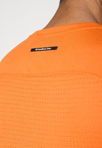 Πορτοκαλί αθλητική μπλούζα με υφασμάτινο σχεδιασμό, που περιλαμβάνει μικρές οπές. Ετικέτα "All Conditions Gear" ραμμένη στο πίσω μέρος του λαιμού.