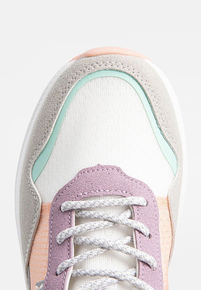 Zapatillas que presentan una parte superior de malla blanca con detalles de ante gris y acentos en verde menta. Superposiciones de ante morado y un detalle texturizado en rosa cerca de los cordones.