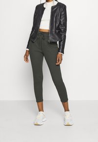 Donna con una giacca di pelle nera, un maglione a collo alto bianco tagliato, pantaloni verde scuro con coulisse e scarpe da ginnastica bianche, in piedi su uno sfondo bianco.