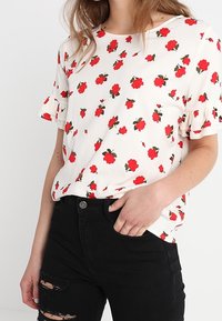Camiseta floral en blanco con patrones de rosas rojas, con mangas cortas con volantes y cuello redondeado. Combinada con jeans negros desgastados.