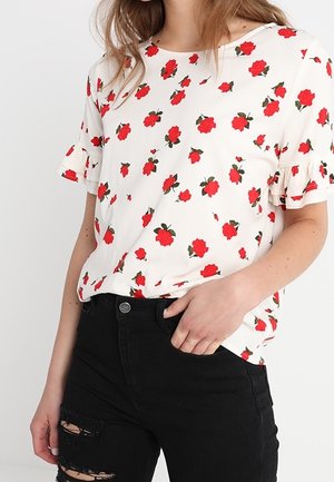 Weißes T-Shirt mit roten Rosenmustern, ausgestattet mit kurzen, gerüschten Ärmeln und einem runden Ausschnitt. Kombiniert mit ausgefransten schwarzen Jeans.