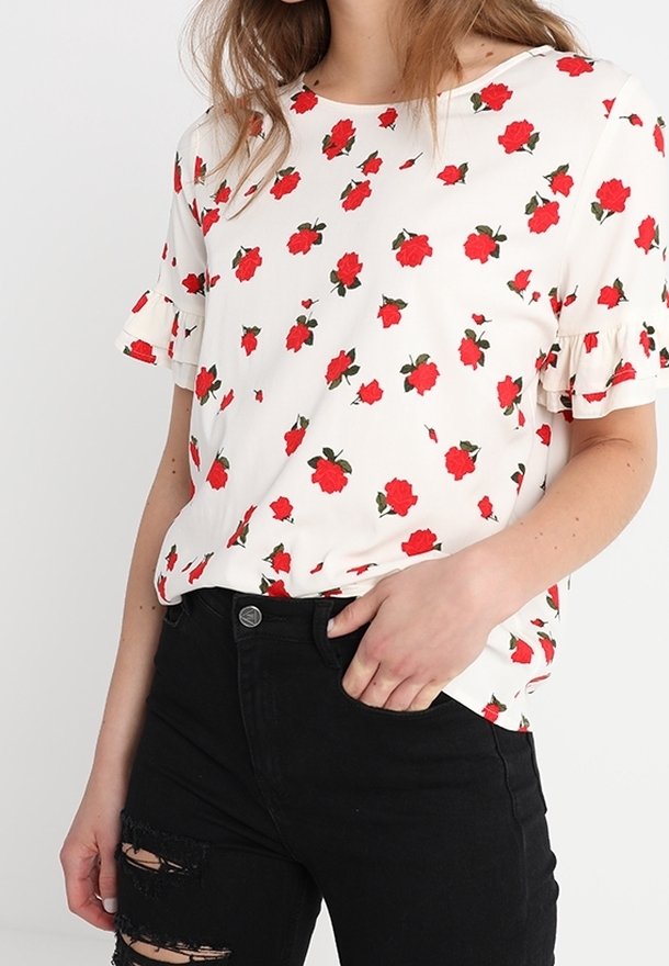 Camiseta floral en blanco con patrones de rosas rojas, con mangas cortas con volantes y cuello redondeado. Combinada con jeans negros desgastados.