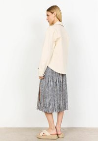 Beige long-sleeve shirt over a patterned midi skirt with blue chevron design, featuring side slits and beige platform sandals.