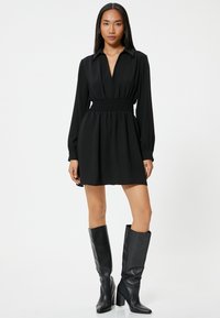 Schwarzes Kleid mit tiefem V-Ausschnitt und gerafftem Taillenbund, langen Ärmeln und fließender Silhouette, kombiniert mit knielangen schwarzen Stiefeln mit Absatz.