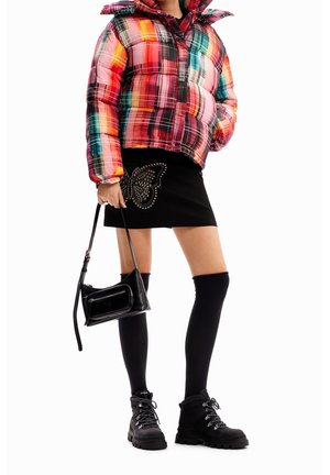 Personne portant une veste matelassée à carreaux multicolores, une jupe noire avec un motif de papillon, des chaussettes montantes noires, des bottes noires, portant un sac à main noir.