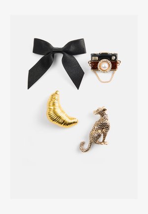 Vier Broschen: eine schwarze Satin-Schleife, eine vergoldete Kamera mit einer Perle, ein goldener Halbmond und ein strukturiertes goldenes Leopardenmotiv mit Punkten.