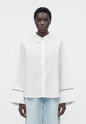 Fekete személy laza fehér, gombos inggel, széles mandzsettával és világoskék farmerrel, egyszínű fehér háttér előtt áll.