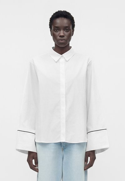 Personne noire portant une chemise blanche ample à boutons avec de larges poignets et un jean bleu clair, debout devant un fond blanc uni.