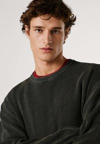 Pull en maille texturée vert foncé avec un col côtelé, superposé à une chemise rouge. Le design présente une coupe ample et des motifs subtils.