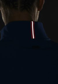 Nära bild av baksidan av en persons nacke som bär en mörk marinblå jacka med en röd och vit vertikal rand på kragen mot en svart bakgrund.