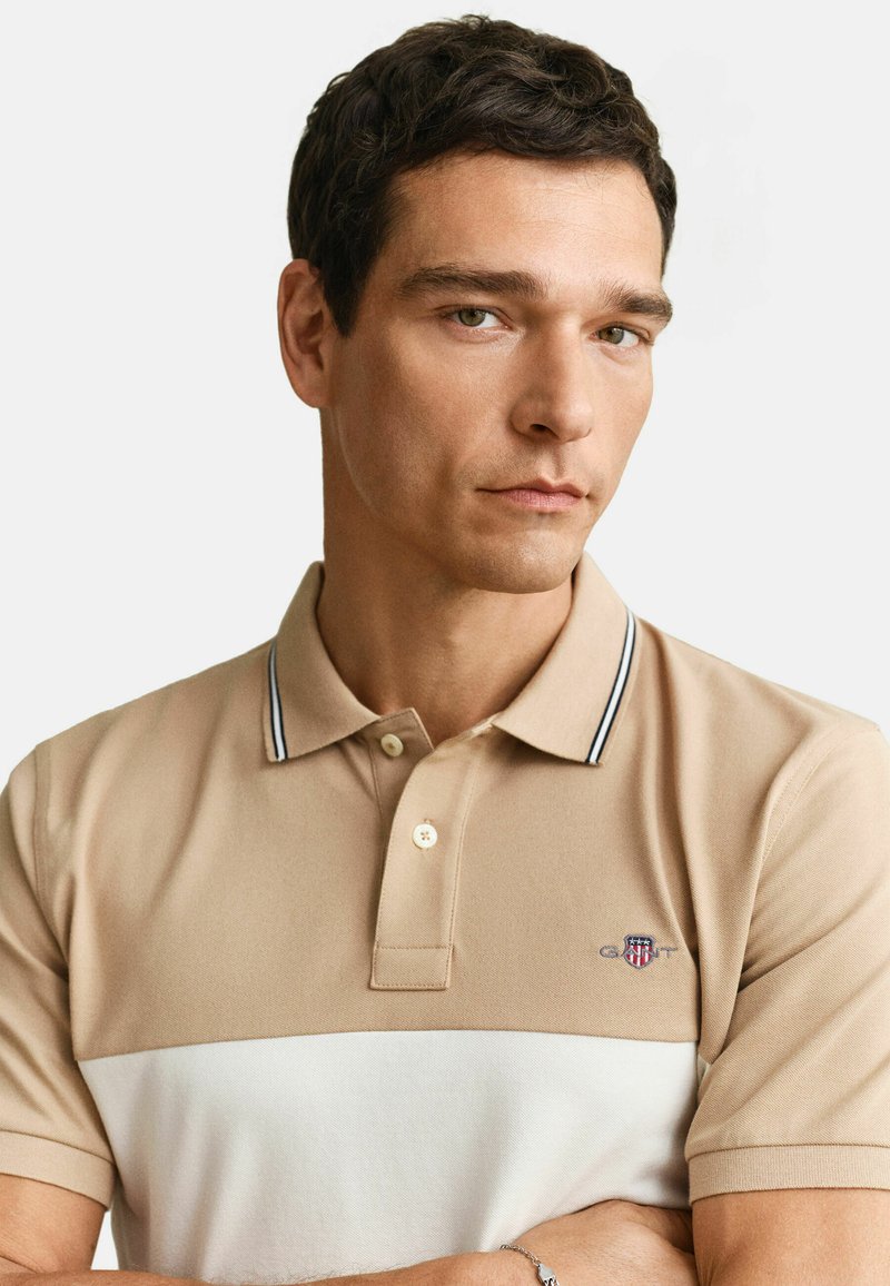 Homme aux cheveux courts et foncés portant un polo beige et blanc avec le logo GANT, regardant légèrement de côté sur un fond uni.