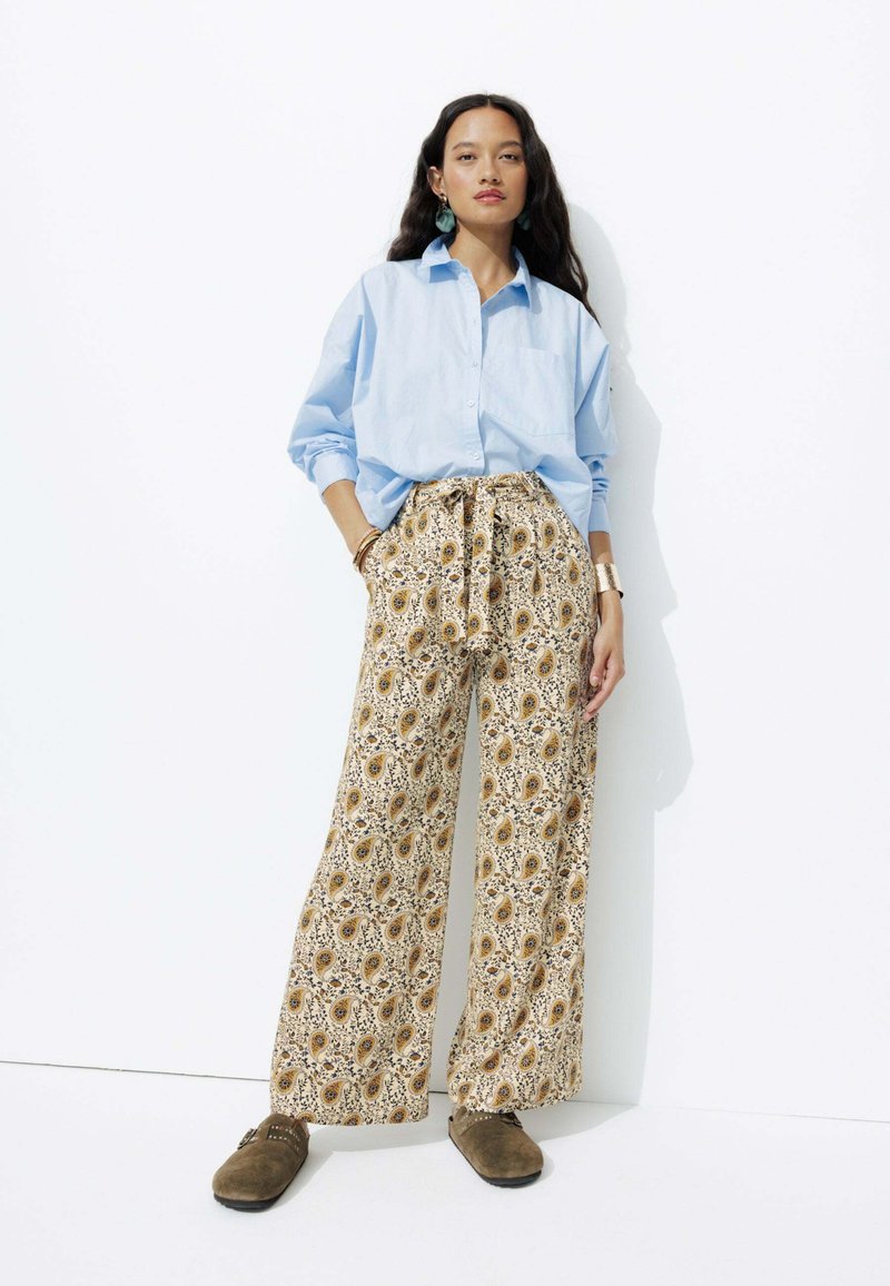 Chemise boutonnée bleu clair, pantalon à jambes larges beige avec un motif en paisley jaune, et chaussures sls en cuir marron.