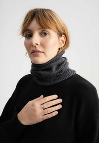 Donkergrijze coltrui met geribbeld textuur, met een gevouwen ontwerp. Zwarte trui eronder, handen op de borst, neutrale achtergrond.