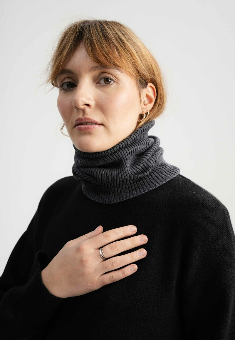 Donkergrijze coltrui met geribbeld textuur, met een gevouwen ontwerp. Zwarte trui eronder, handen op de borst, neutrale achtergrond.