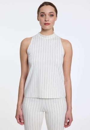 Vrouw die een witte mouwloze top en bijpassende broek met dunne zwarte verticale strepen draagt, kijkt recht vooruit met een neutrale uitdrukking.