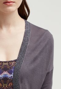 Grå cardigan med en glittrande silverkant längs halslinjen. Texturen verkar mjuk, med en lagerdesign över en mönstrad topp under.