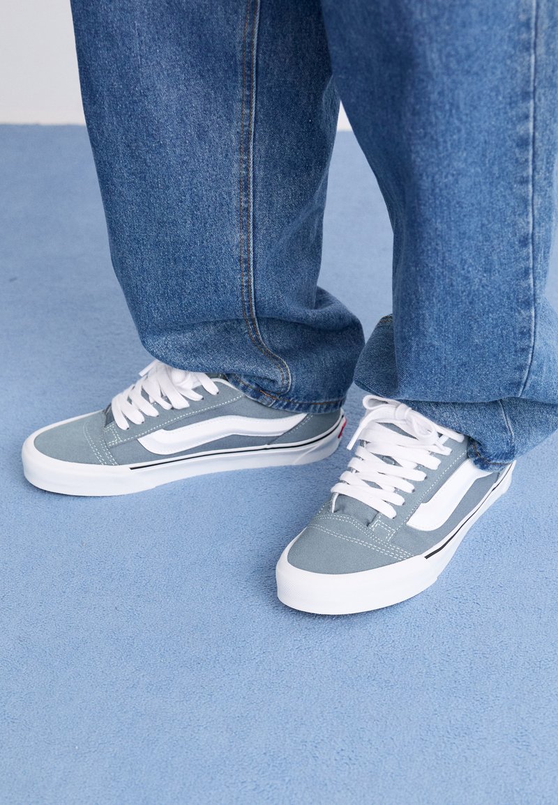 Persona que lleva zapatillas ligeras de color azul y blanco con cordones blancos y jeans sueltos de color azul, de pie sobre un suelo alfombrado de color azul claro.