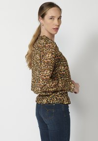 Blusa nera trasparente con motivo floreale in giallo, arancione e rosa. Presenta un colletto arricciato e un orlo smock, abbinata a jeans blu scuro.
