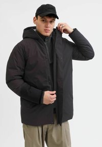Sort puffjakke med hætte, lynlås og trykknapper; med et struktureret inderlag og parret med en strikket sweater under.