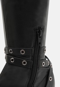 Botte en cuir noire montante avec une fermeture éclair latérale, des sangles décoratives et des œillets en métal, présentant une texture lisse et un design élégant.