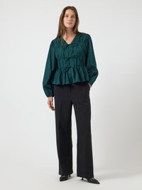 Blusa verde a scollo a V con dettagli arricciati e lacci sul davanti, abbinata a pantaloni neri a vita alta e décolleté neri a punta.