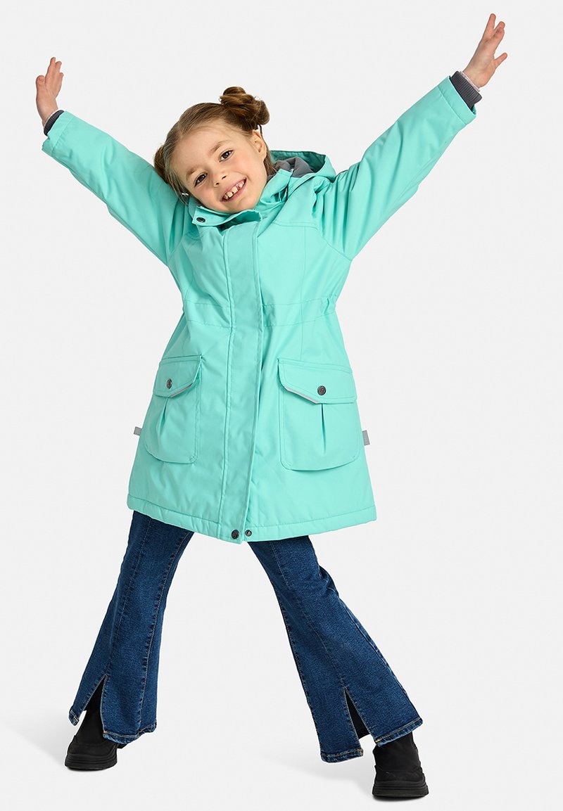 Šviesiai mėlyna atspari vandeniui striukė su gaubtu, dviem priekinėmis kišenėmis ir surišta liemeniu, derinama su tamsiai džinsinėmis platėjančiomis kelnėmis ir juodais batais.