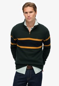 Zielony sweter z dzianiny z suwakiem na ćwierć, z poziomymi paskami w granatowym i musztardowym kolorze. Nosi się go na jasnoszarym pasekowym t-shircie oraz ciemnych dżinsach.