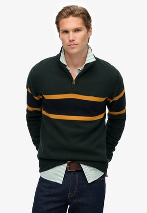 Grøn strikket pullover med lynlås halvlukket, med vandrette striber i marineblå og sennep. Båret over en lys blå stribet skjorte og mørke jeans.