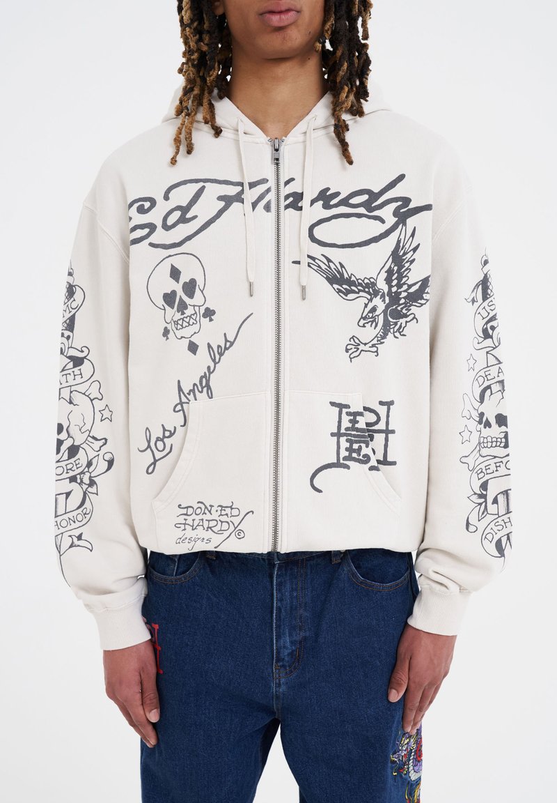 Sudadera con cremallera color beige claro con estampados gráficos en negro, incluyendo calaveras y un águila. Cuenta con bolsillos frontales y puños acanalados.