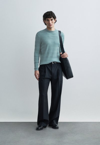 Φωτεινό γαλάζιο πλεκτό πουλόβερ με ριγέ υφή, σκούρο πλισέ παντελόνι και μαύρη δερμάτινη τσάντα ώμου. Το μοντέλο στέκεται μπροστά σε μονοχρωμο φόντο.