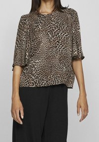 Blusa con stampa leopardata, colletto rotondo, maniche corte e vestibilità comoda; realizzata in tessuto morbido con una texture setosa. Pantaloni neri visibili sotto.