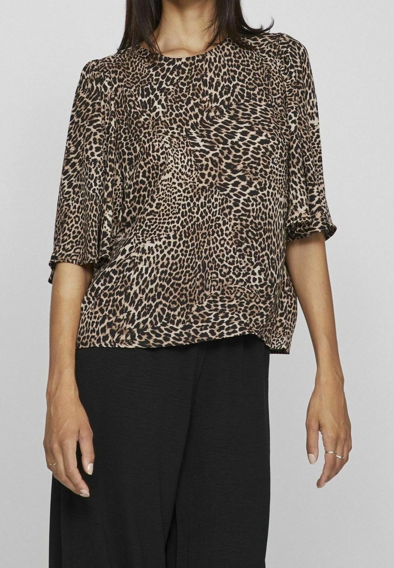 Blusa con stampa leopardata, colletto rotondo, maniche corte e vestibilità comoda; realizzata in tessuto morbido con una texture setosa. Pantaloni neri visibili sotto.