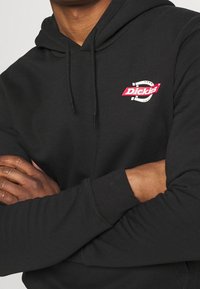 Svart hoodie i bomull med en frontlogo i rött och vitt. Detaljer inkluderar en känguruficka och en justerbar huva.