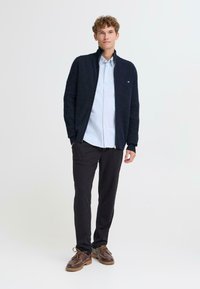 Námořnický žebrovaný cardigan s límečkem přes světle modrou košili s knoflíky. Černé kalhoty a hnědé lodní boty doplňují celkový vzhled.