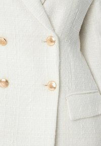 Bílý texturovaný blazer se zapínáním na dvě řady knoflíků, zlatými knoflíky a jednou kapsou na přední straně.