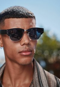 Ochelari de soare negri cu un accent metalic, având o formă rectangulară și lentile închise la culoare. Purtați de o persoană cu părul scurt și pielea clară.