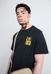 Zwarte katoenen t-shirt met een gele Timberland-logo patch op de borst. Model draagt een beige schoudertas met een rits.