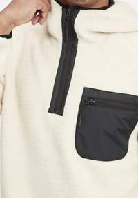 Crème kleurige fleece trui met zwarte ritszak en details, een halslijn met halve rits, een gestructureerd oppervlak en een capuchon.