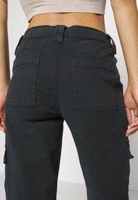 Femme portant un pantalon cargo noir avec de grandes poches arrière, montrée de la taille jusqu'à mi-cuisse, sur un fond clair uni.