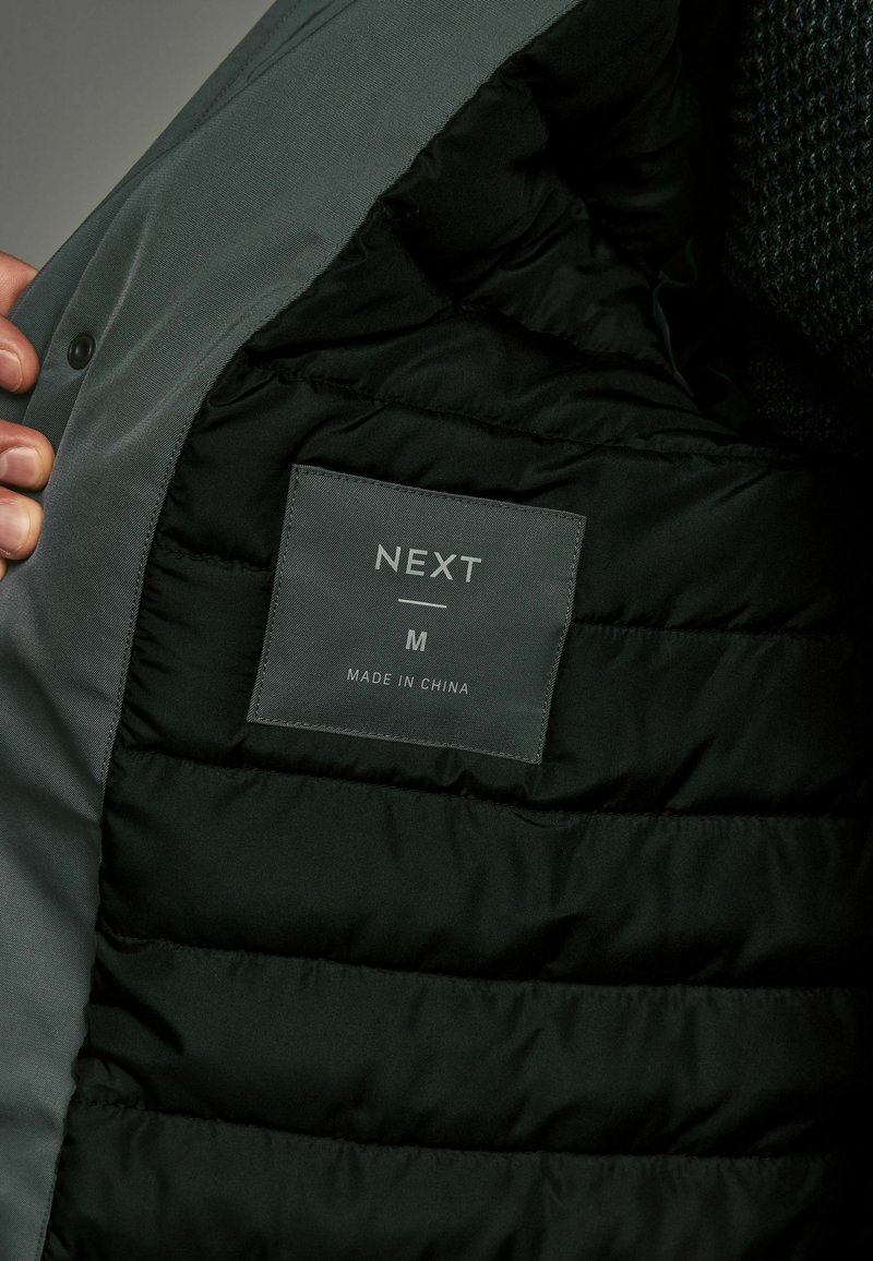Interior de chaqueta tipo acolchado gris con forro negro acolchado. La etiqueta dice "NEXT", talla M, indicando que es de origen chino. Presenta cierre a presión.