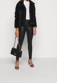 Chaqueta de piel sintética negra, blusa blanca con detalles de encaje, leggings negros brillantes, bolso negro con herrajes dorados y tacones negros con tiras.