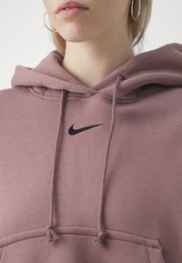Huppari hillityssä malvan sävyssä, pehmeästä kankaasta valmistettu, jossa on kiristysnyörit ja rinnassa musta Nike-logo kirjailtuna.