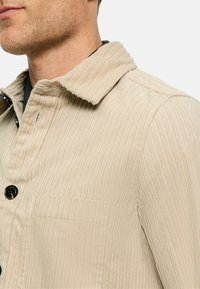 Beige strukturierte Jacke mit vertikalen Rippungen, einem umgeschlagenen Kragen und schwarzen Knöpfen. Verfügt über eine Brusttasche und ein lässiges Design.