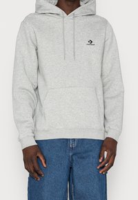 Sudadera con capucha gris hecha de tela suave y texturizada. Cuenta con un bolsillo frontal y una capucha con cordón ajustable. Logo de Converse en negro en el pecho.