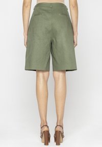 Oliwkowe lniane szorty bermudy o luźnym kroju, podkreślone średnim stanem i dwiema tylnymi kieszeniami. Na nogach znajdują się masywne brązowe sandały.