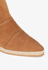 Botín de ante en marrón claro con punta afilada. Presenta un ribete trenzado de yute a lo largo de la suela, que resalta su superficie texturizada.